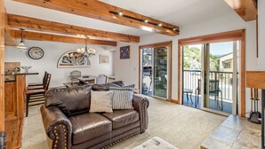 A spacious living room with a leather sofa, wooden beams, a chandelier, and a view through glass doors to an outdoor patio. - Charming 2BR/2BA Snowmass Condo with Shared Hot Tub (Snowmass Village)