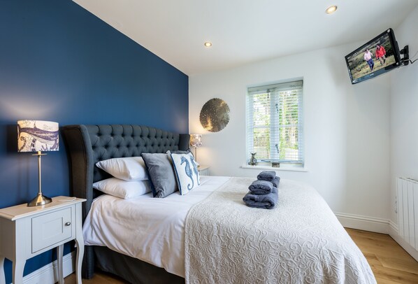 A bedroom with a bed, bedside table, lamp, wall-mounted TV, and a window with blinds.