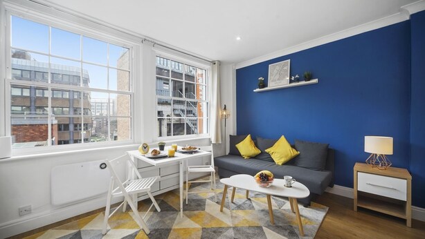 A modern living room with a white sofa, a dining table with chairs, a coffee table, and a blue accent wall. - Studio Apartment Blue (London)