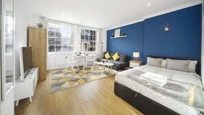 A modern hotel room with a large bed, a desk with a chair, a sofa, and a small table with a lamp. - Studio Apartment Blue (London)