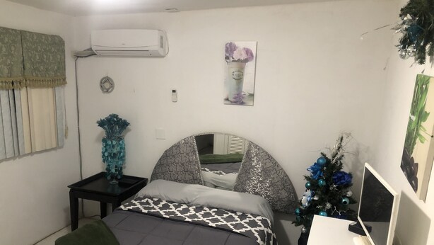 A bedroom with a bed, a desk with a computer, an air conditioner, a small table with a vase, and a wall-mounted clock.