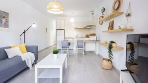 Ein modernes Wohnzimmer mit grauem Sofa, weißem Couchtisch und Essbereich mit weißem Tisch und Stühlen. Holzregale mit Dekoration und Pflanzen, und eine Küche im Hintergrund.