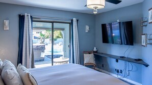 A hotel room with a bed, a ceiling fan, a TV mounted on the wall, and a view of an outdoor swimming pool. - Oasis Home with Pool and Hot Tub (Lake Havasu City)