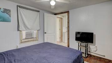 Una camera da letto con un letto, un ventilatore a soffitto, una finestra con persiane, una televisione e una porta che dà su un'altra stanza.