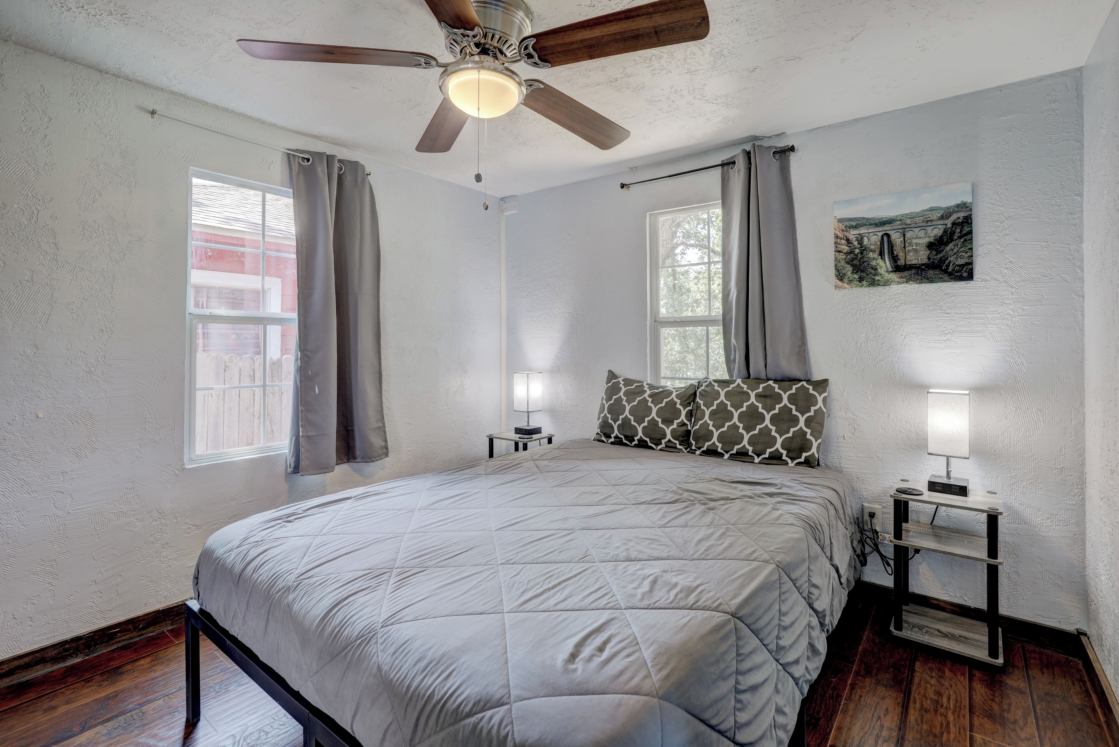 A bedroom with a bed, a ceiling fan, a nightstand with a lamp, and a painting on the wall.