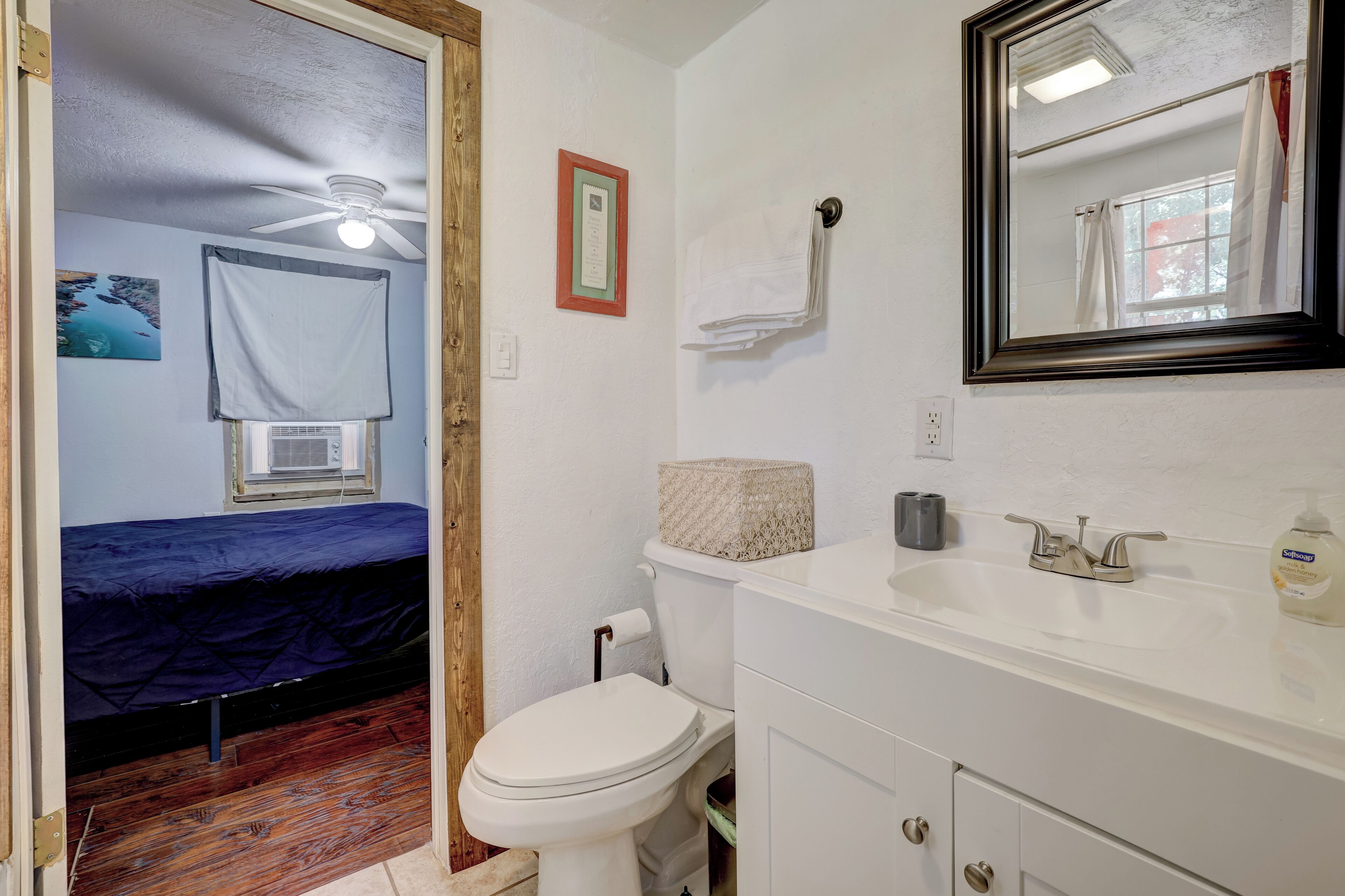A small, compact bathroom with a white sink, toilet, and a mirror above the sink. There is a bed with a blue mattress in the adjacent room.