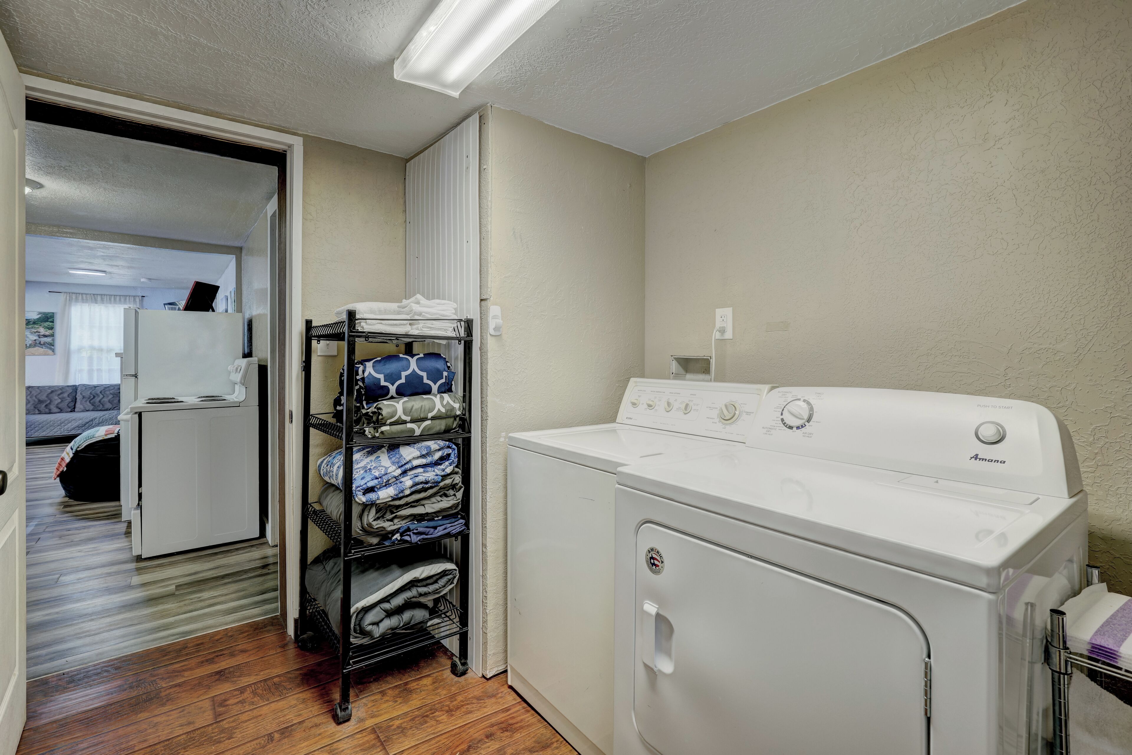 A laundry room with a white washer and dryer, a shelf with folded towels, and a door leading to a bedroom.