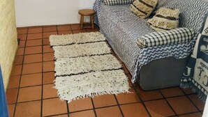 Een kleine, knusse kamer met een geruit sofa, een klein krukje en een vloerkleed met patroon op terracotta tegels.