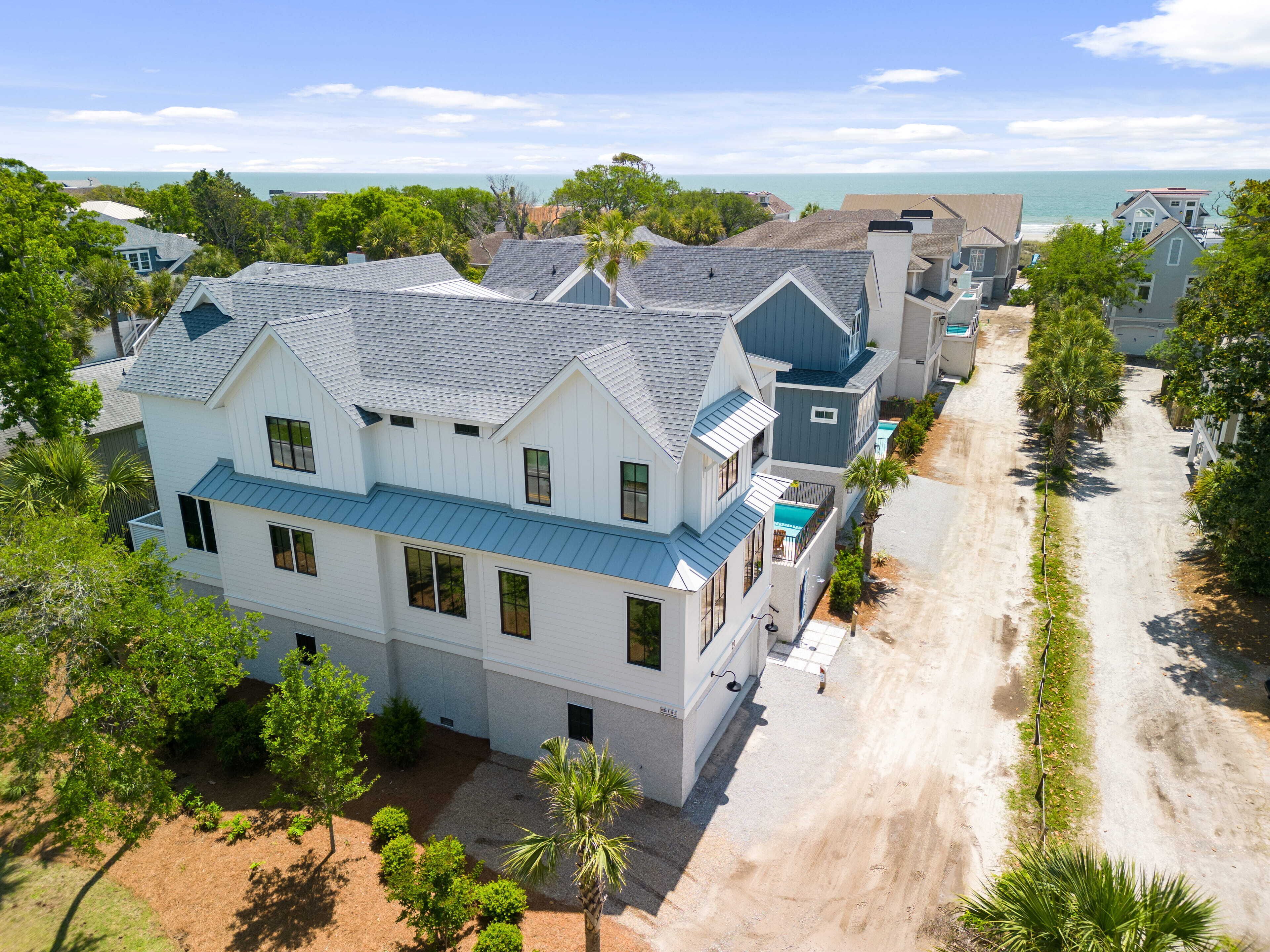 A coastal residential area with a large house featuring a blue roof and white walls, surrounded by trees and a sandy path.