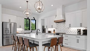 Cocina moderna con armarios blancos, una isla grande con taburetes de bar, electrodomésticos de acero inoxidable e iluminación colgante.