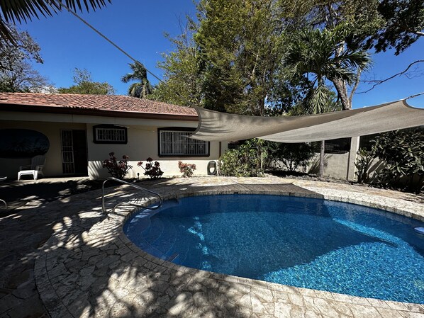A swimming pool with a tiled deck, surrounded by a house with a tiled roof and a shaded area.