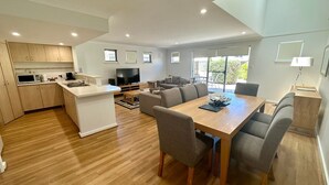 A modern kitchen with a central island, a dining area with a wooden table and grey chairs, and a living area with a sofa and TV.