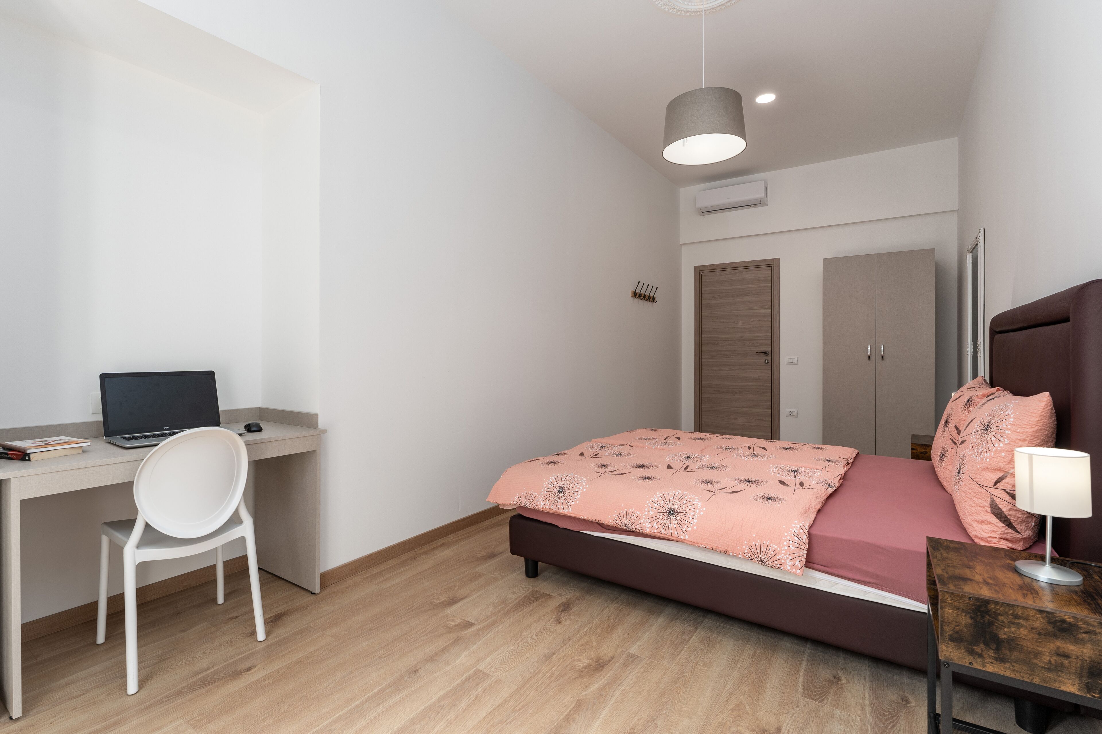 A bedroom with a bed, a desk with a laptop, a chair, a bedside table with a lamp, and a wardrobe.