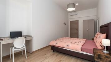 A bedroom with a bed, a desk with a laptop, a chair, a bedside table with a lamp, and a wardrobe.
