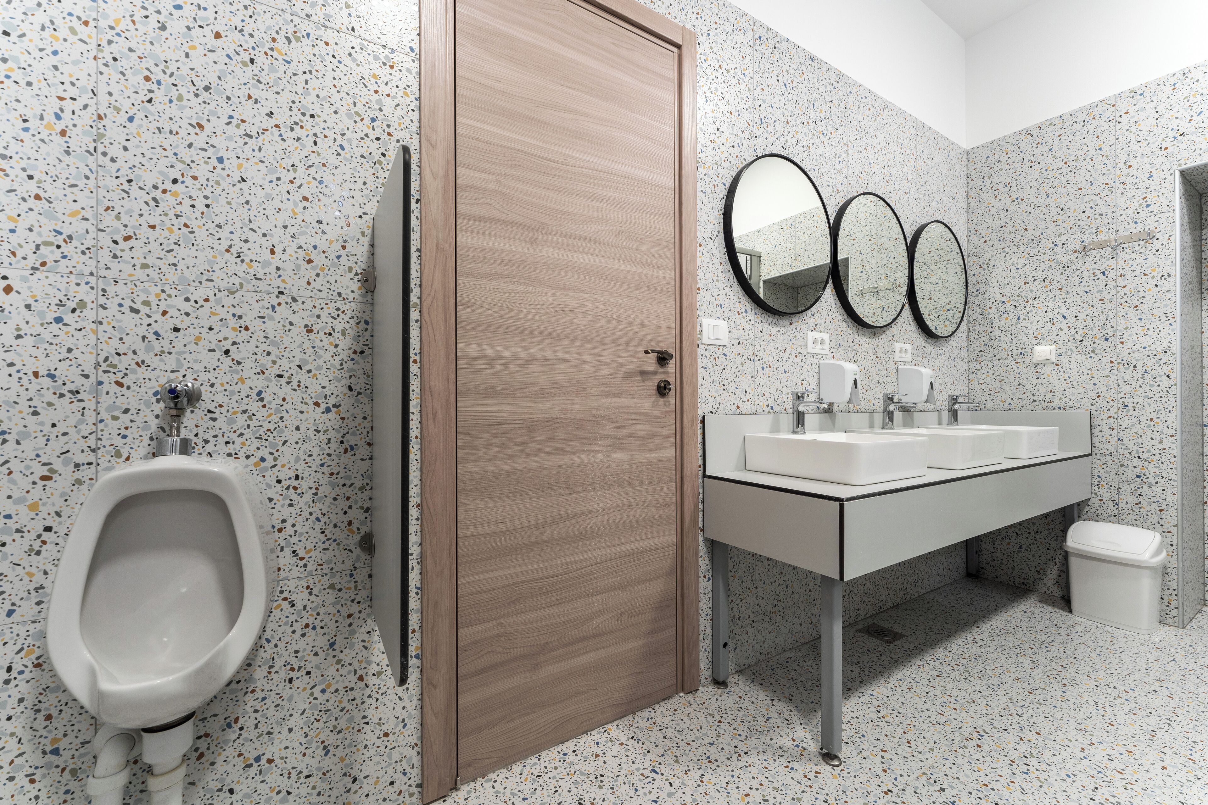 A modern bathroom with a urinal, a wooden door, and a white sink with two mirrors.