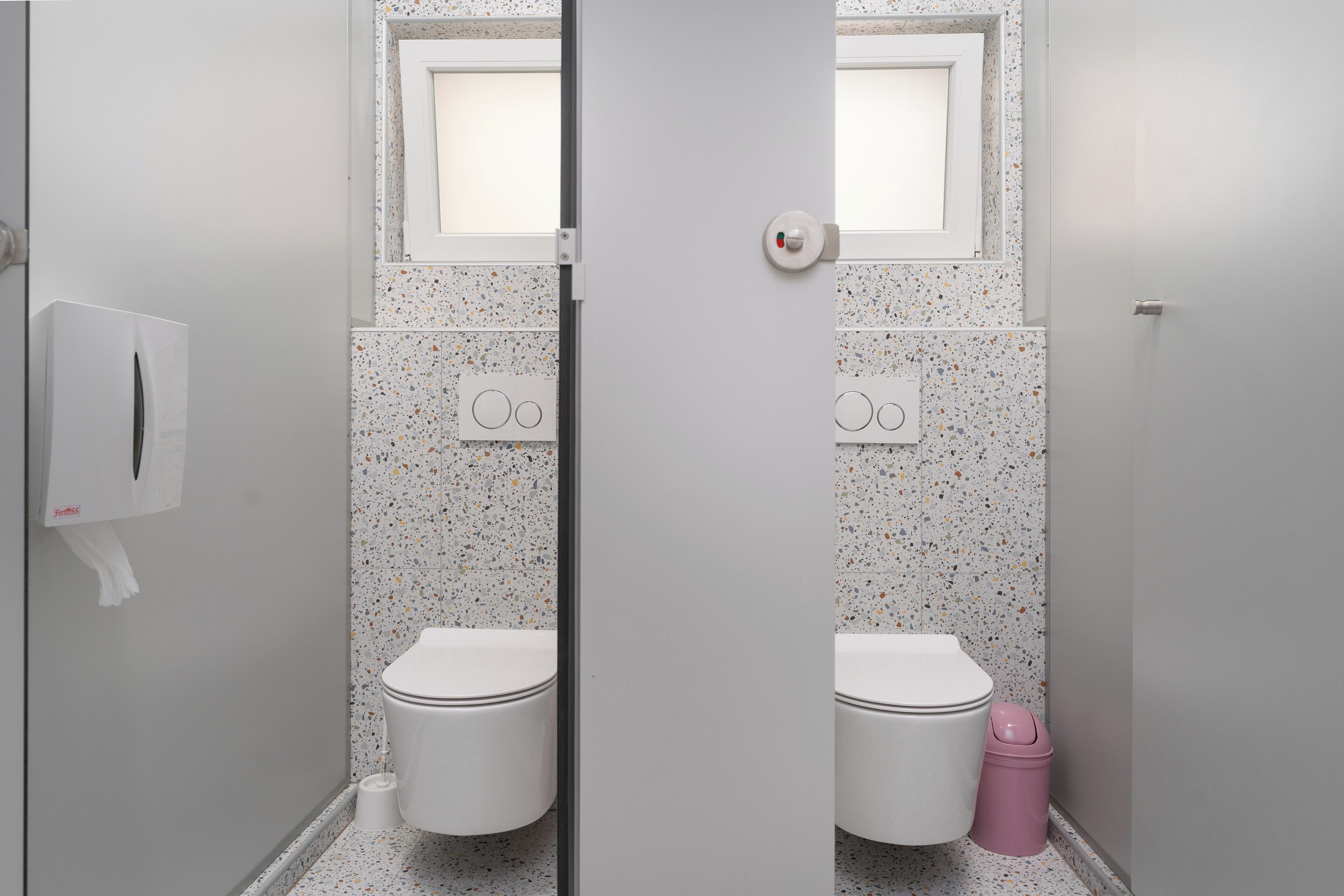 A bathroom with two toilets, each with a small window above. There is a trash bin next to the right toilet.