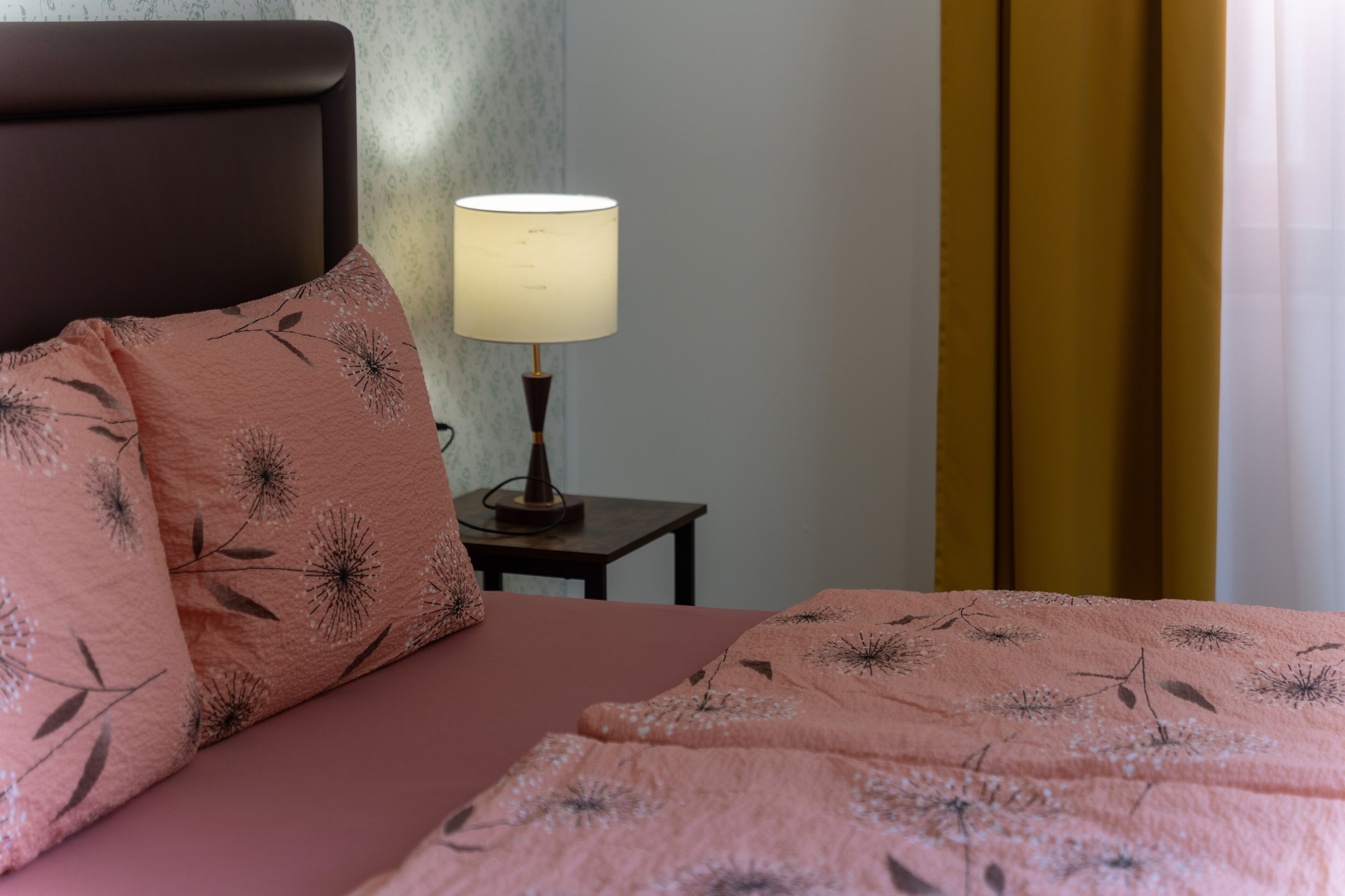 A neatly made bed with a pink floral bedspread and matching pillows, a bedside lamp, and a small wooden nightstand.