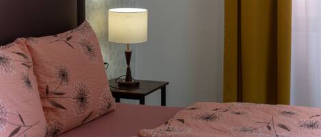 A neatly made bed with a pink floral bedspread and matching pillows, a bedside lamp, and a small wooden nightstand.