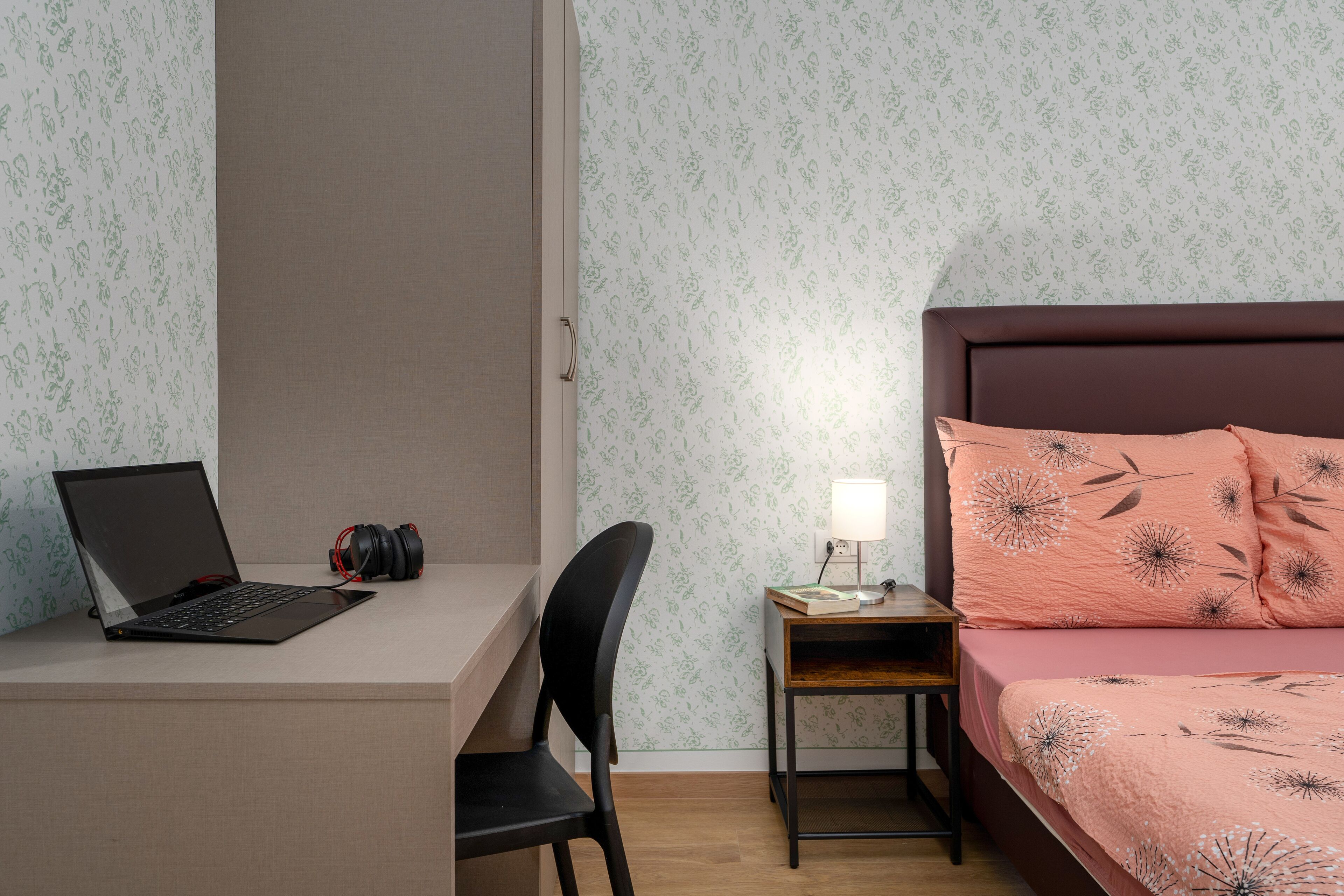 A bedroom with a bed, a desk with a laptop and headphones, a chair, and a bedside table with a lamp.
