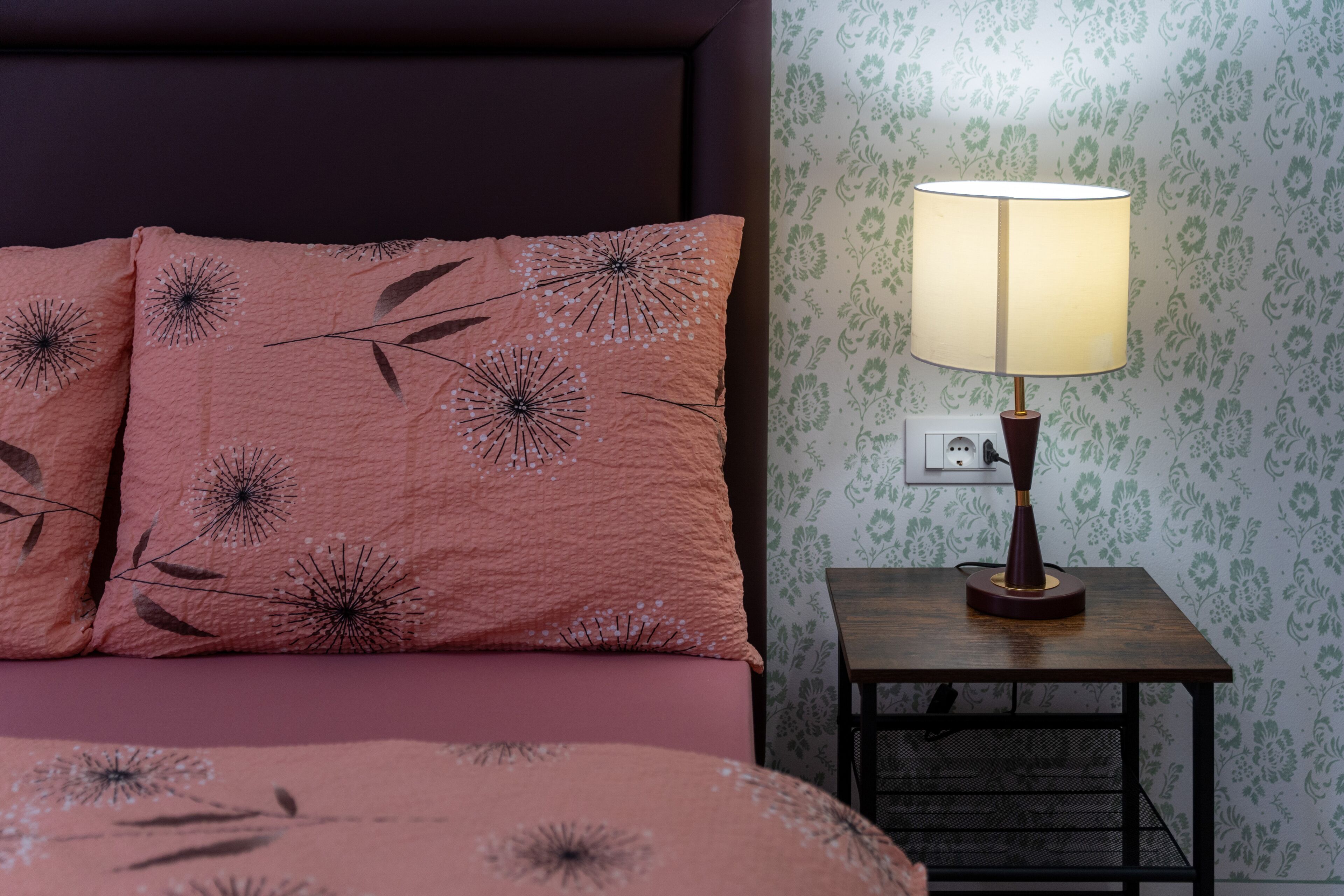 Ein ordentlich bezogenes Bett mit rosa Bettwäsche und einem blumigen Kissen, ein Nachttisch mit Lampe sowie eine Wand mit floralem Tapetenmuster.