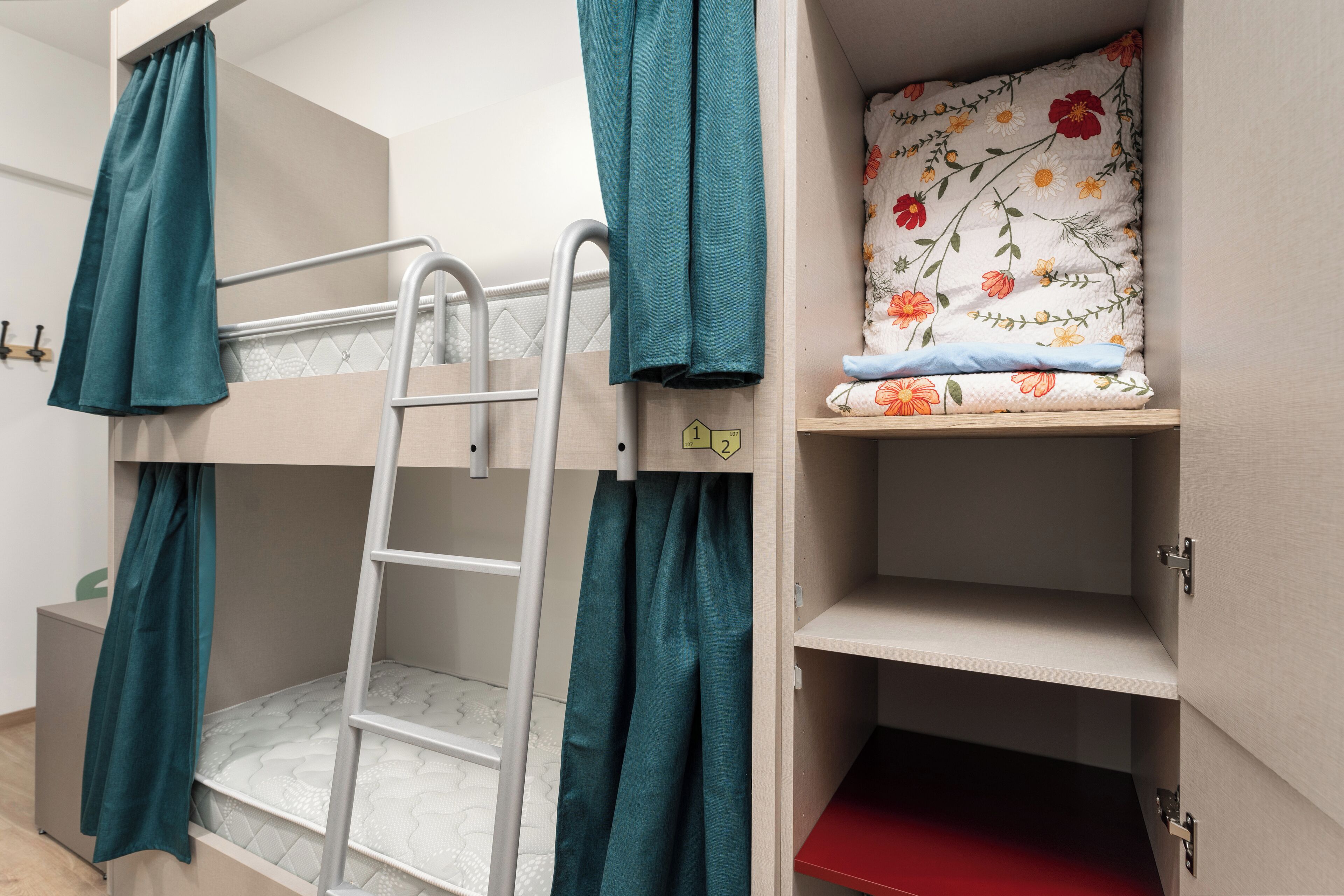 A bunk bed with a ladder, teal curtains, and a floral-patterned pillow on a shelf.