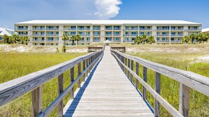 A wooden boardwalk leading to a multi-story hotel with balconies, surrounded by grassy areas and palm trees. - SunTime at the BEACH! Updated 2nd Floor Unit! GREAT Views of 2 Pools and Gulf! (Dauphin Island)