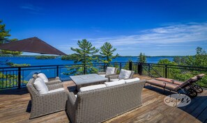 A deck with wicker furniture, a table, and an umbrella, overlooking a lake with trees and a clear sky. - Incredible 9 bdrm, 9 bath, Tennis Court! Nanny Suite! Gym! Lake Muskoka!!! (Port Carling)