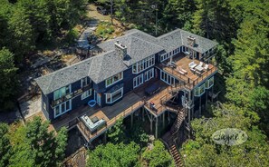 A large, multi-story house with a prominent deck area, surrounded by dense forest. - Incredible 9 bdrm, 9 bath, Tennis Court! Nanny Suite! Gym! Lake Muskoka!!! (Port Carling)