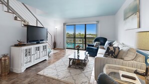 A living room with a TV on a white cabinet, a sofa, a coffee table, and a view of the outdoors through a glass door. - Beach Walk 18 Cozy Condo-Walk to Hangout Festival - Private Pool, Free Parking (Gulf Shores)