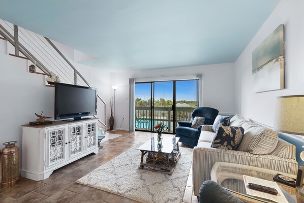 A living room with a TV on a white cabinet, a sofa, a coffee table, and a view of the outdoors through a glass door.