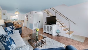 A modern living room with a sofa, coffee table, TV, and staircase. - Beach Walk 18 Cozy Condo-Walk to Hangout Festival - Private Pool, Free Parking (Gulf Shores)