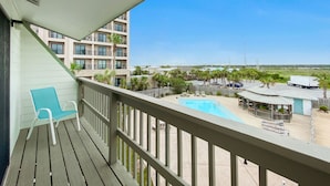 A balcony with a blue chair, a pool area with lounge chairs, a gazebo, and a view of a multi-story building in the background. - Beach Walk 18 Cozy Condo-Walk to Hangout Festival - Private Pool, Free Parking (Gulf Shores)