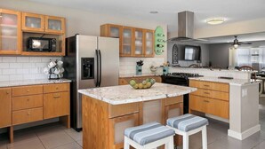 Una cocina moderna con armarios de madera, un refrigerador de acero inoxidable, un microondas y una estufa. Hay una pequeña isla con taburetes y un cuenco con fruta.