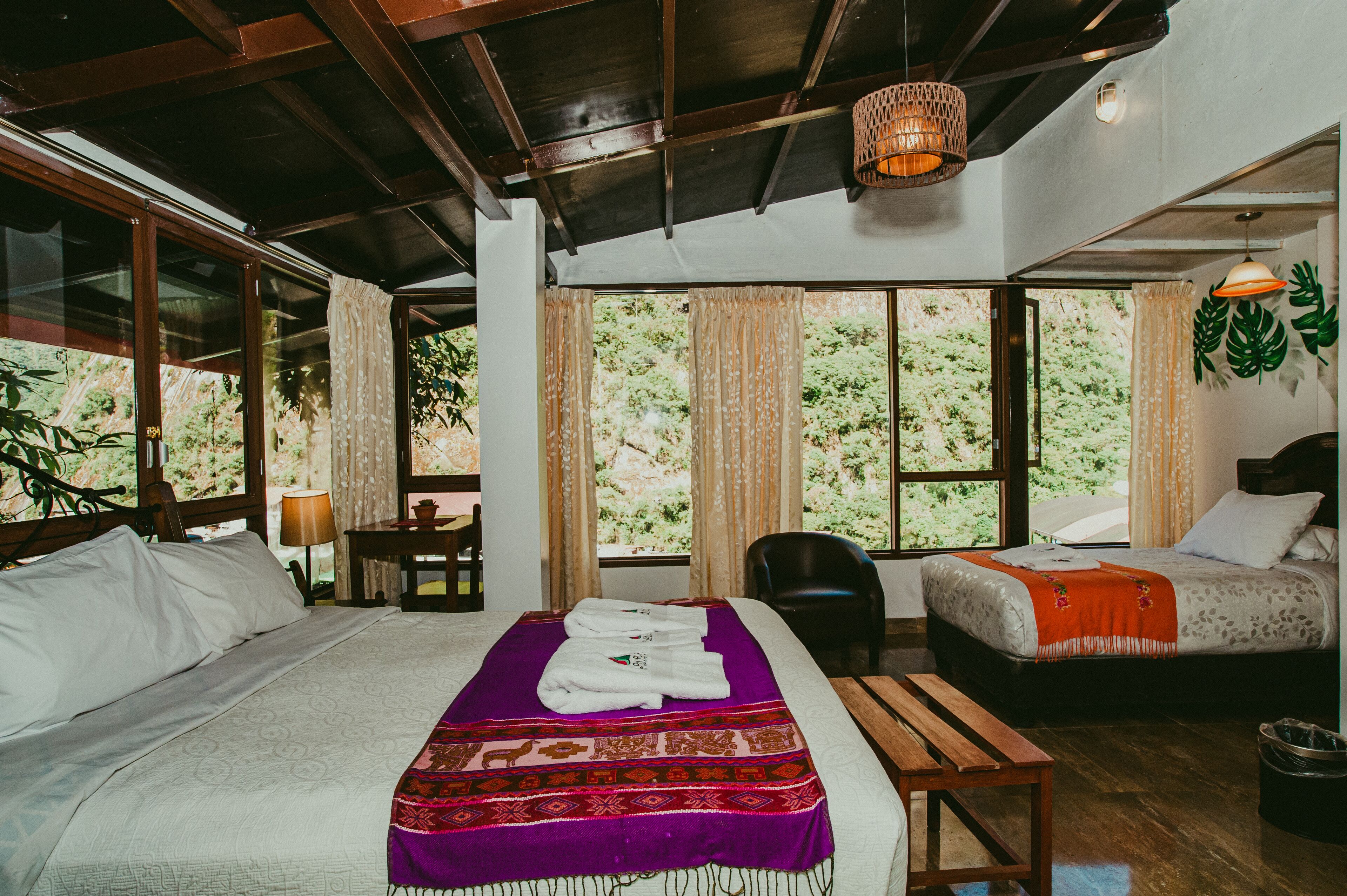 A room with a large bed, a smaller bed, a desk, and a bench. There are curtains and a view of greenery outside.