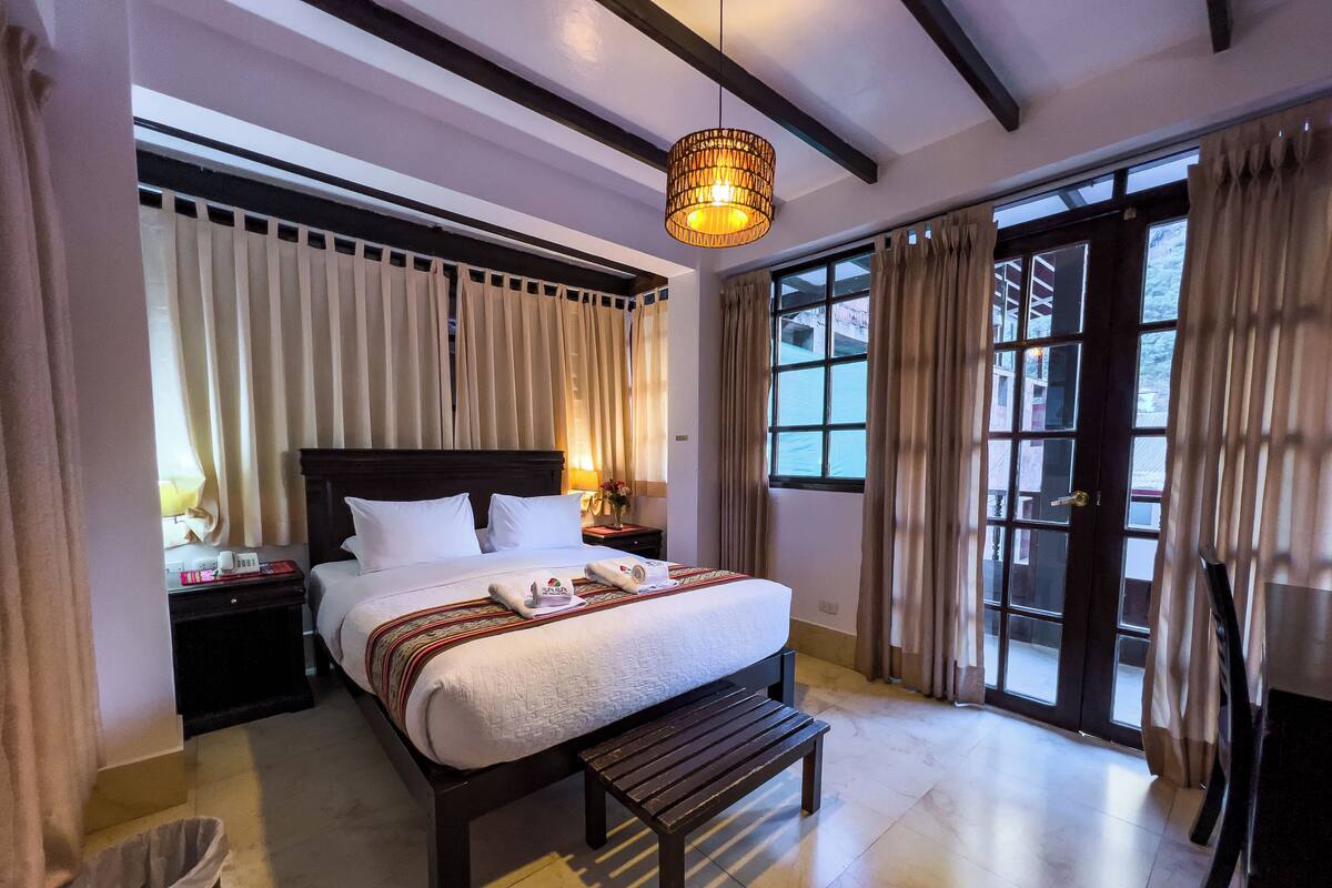 A hotel room with a large bed, a bedside table with a lamp, a bench, and a view of the outside through large windows.