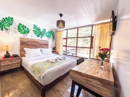 Un dormitorio bien iluminado con una cama de madera, una mesita de noche con una lámpara, un escritorio con un florero y un gran ventanal con cortinas.