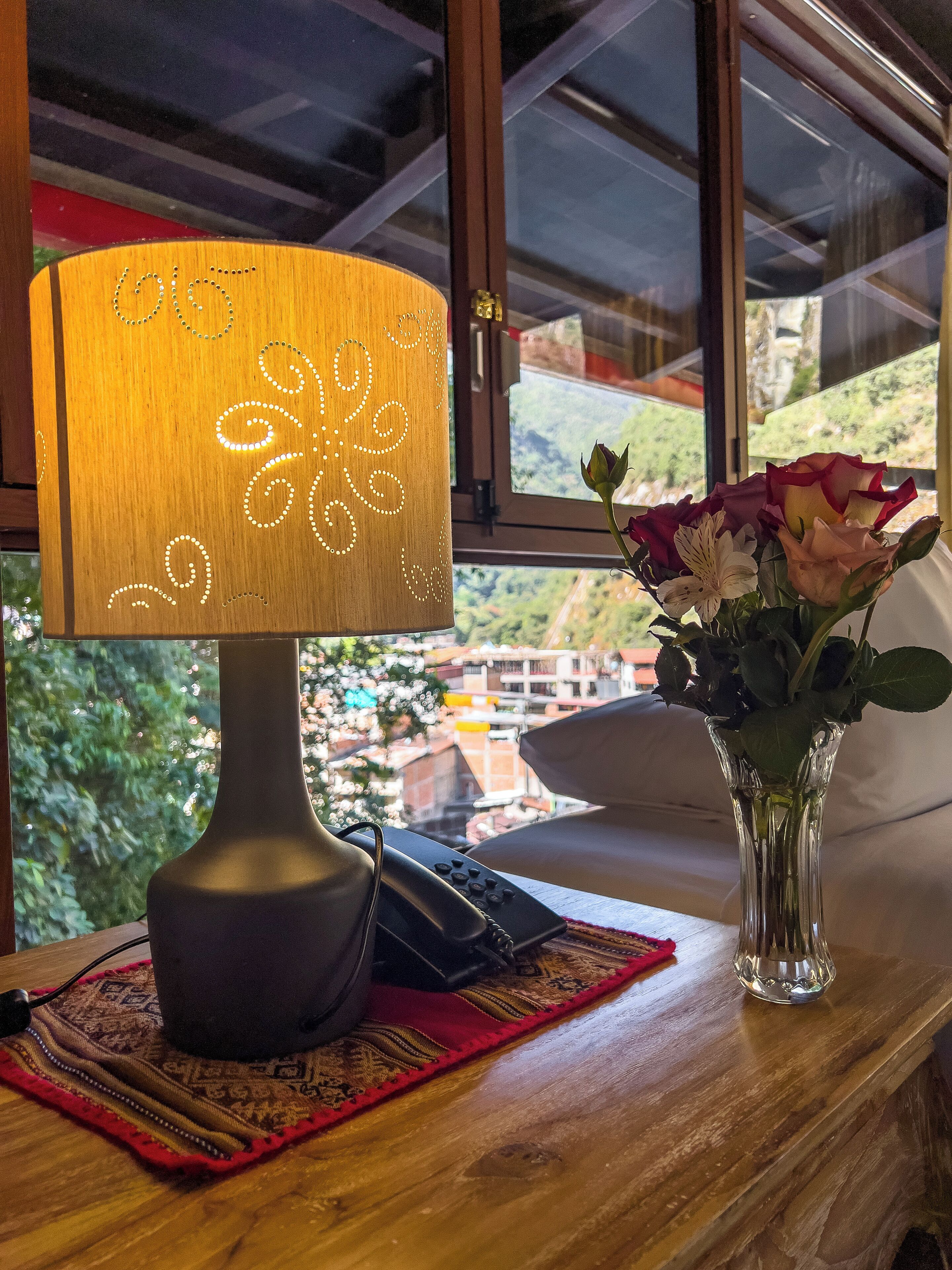En lampa med en lampskärm med mönster, en vas med blommor och en utsikt över ett bostadsområde genom ett fönster.