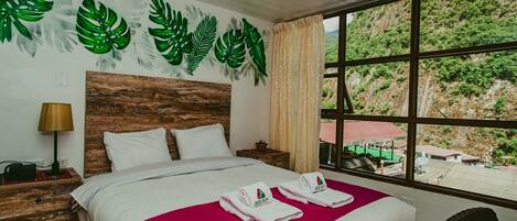 A hotel room with a bed, a nightstand, a lamp, a window with a view of greenery, and a wall with leaf decorations.
