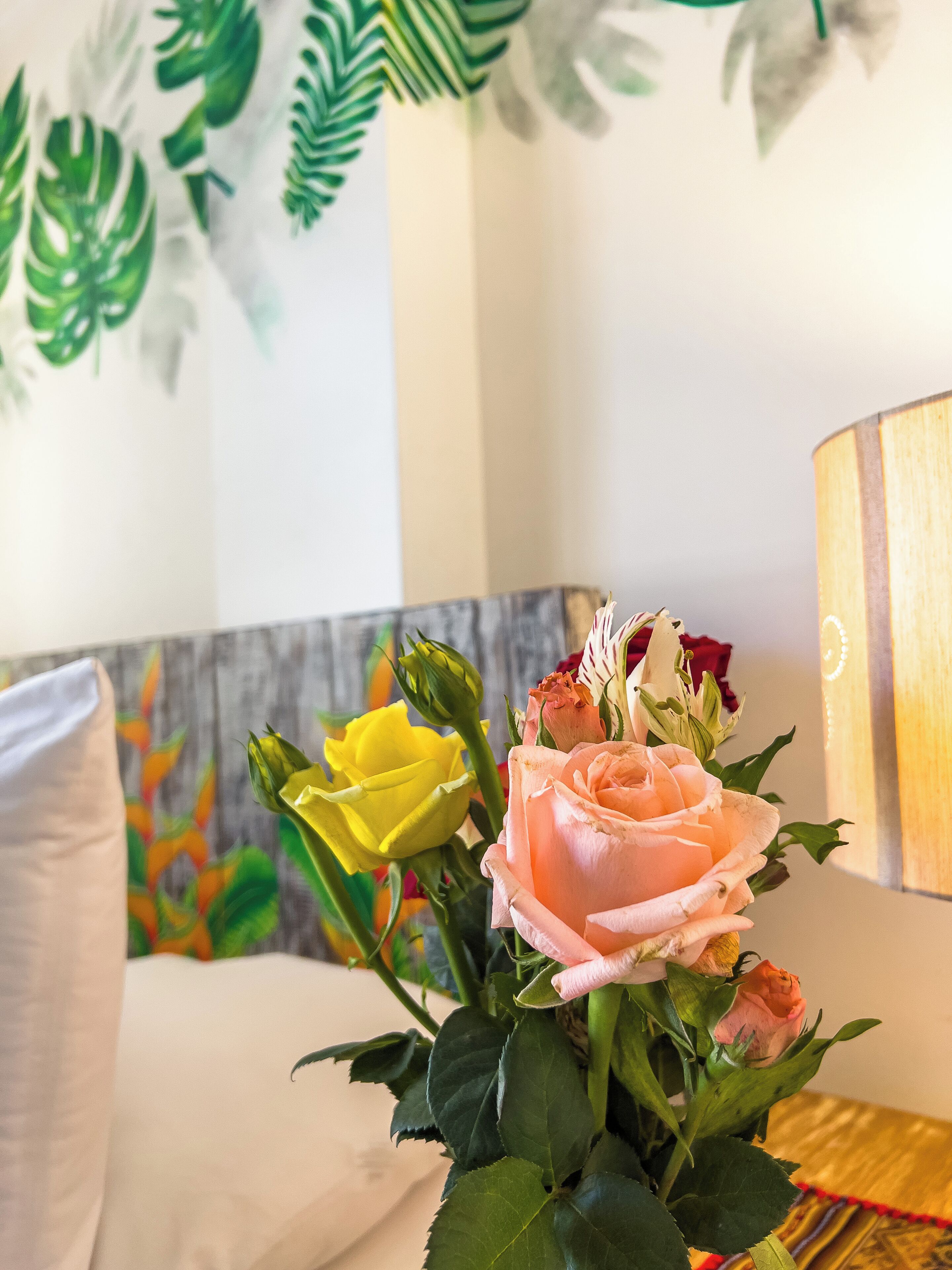 Een kamer met een bed, een muur met een groene bladeren muurschildering, en een vaas met rozen.