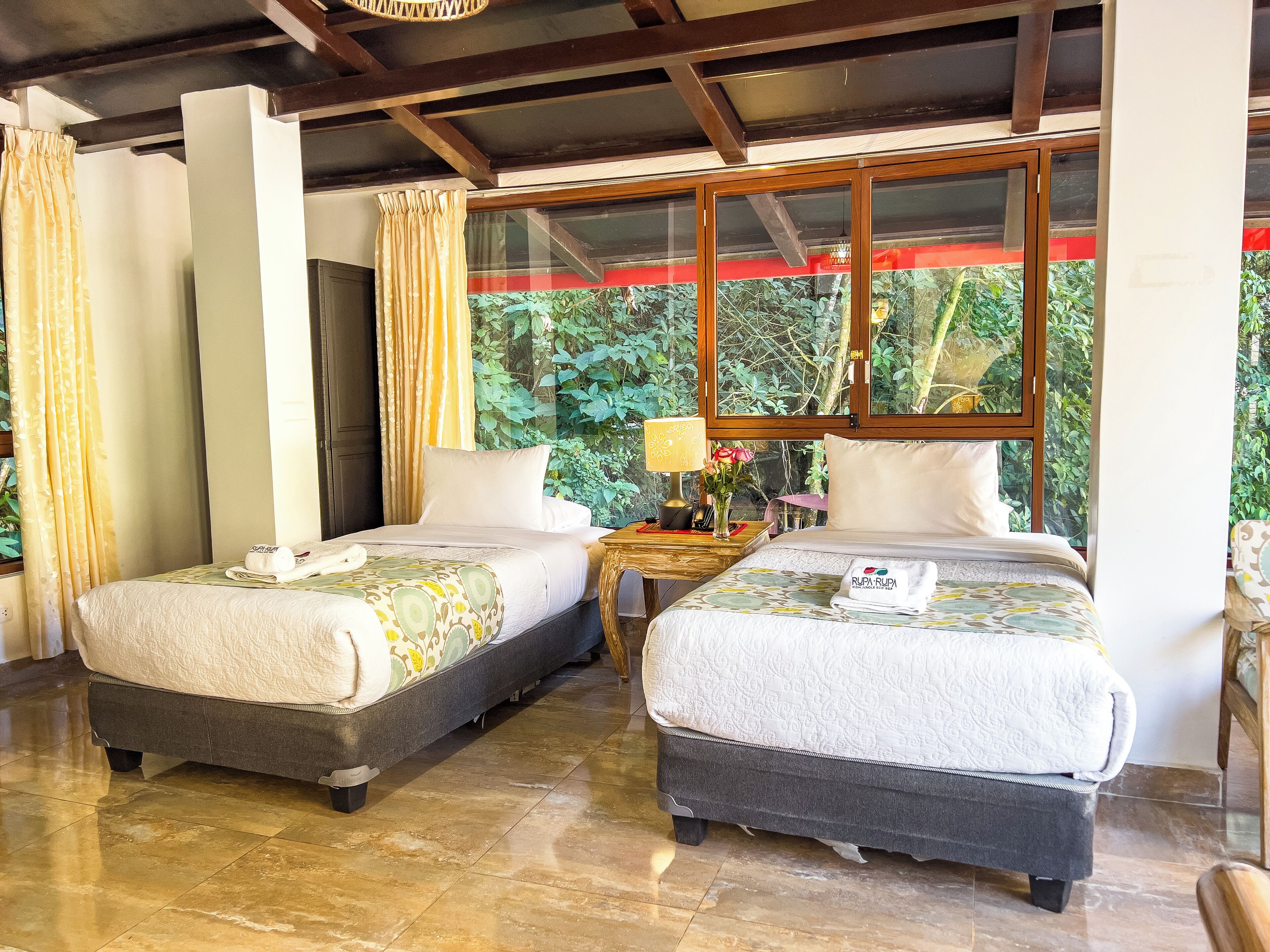 Habitación de hotel con dos camas, techo de madera, ventana con vistas a la vegetación y una mesita con lámpara y flores.