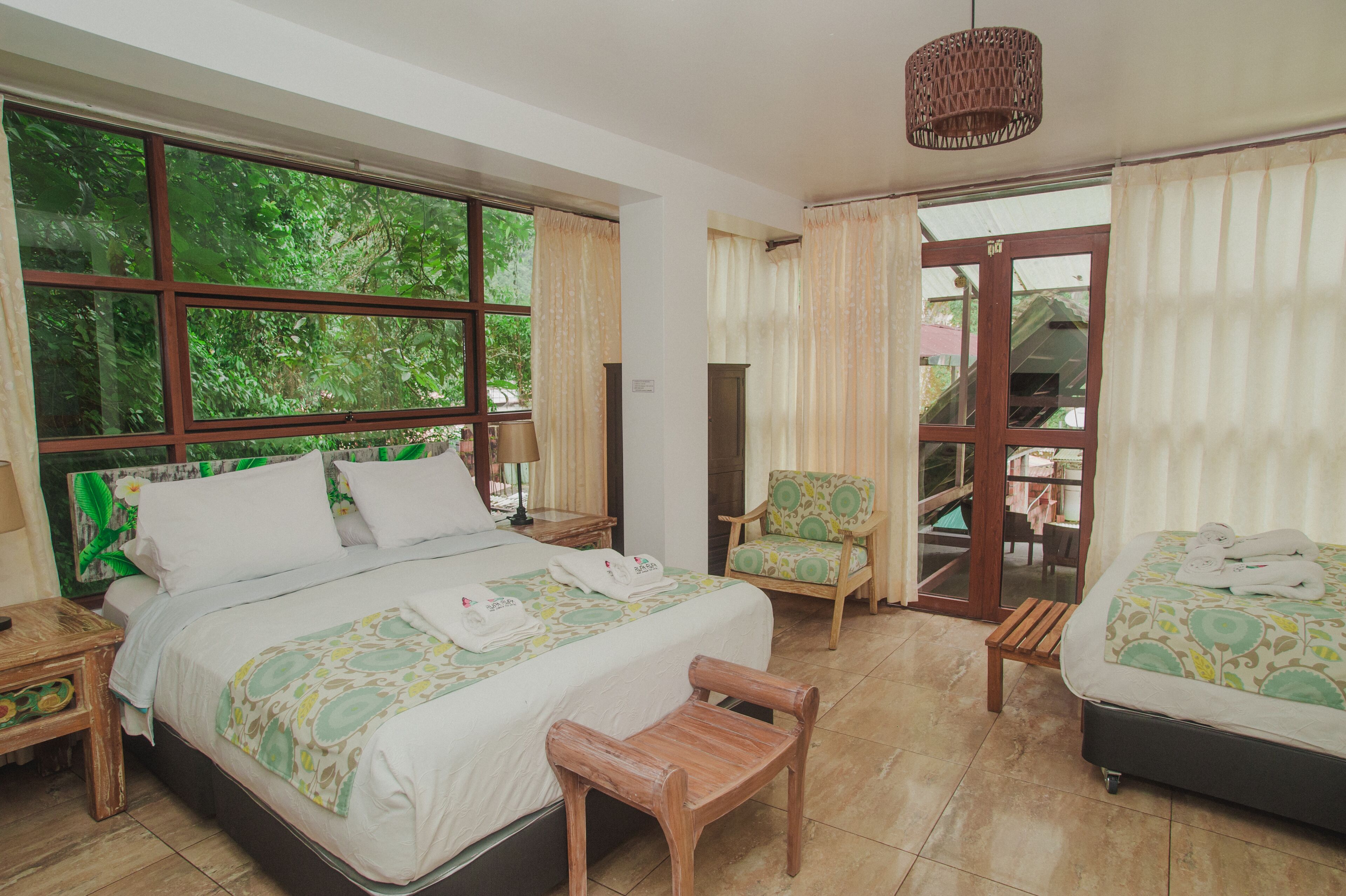 Quarto de hotel com uma cama grande, uma cama menor, uma cadeira, um banco e vista para o verde pelas janelas.