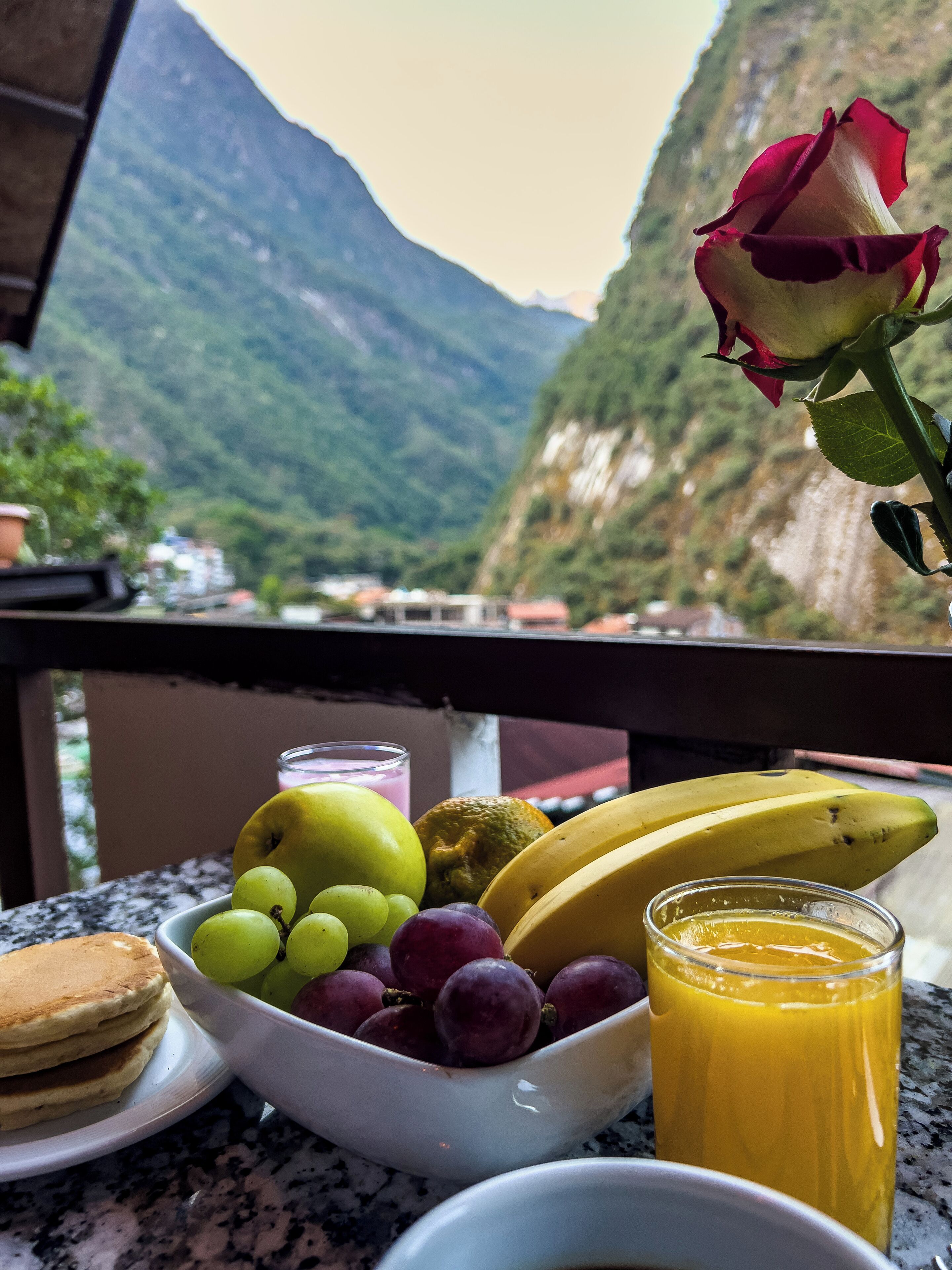 A table with a fruit bowl, a glass of orange juice, a plate of pancakes, and a rose.