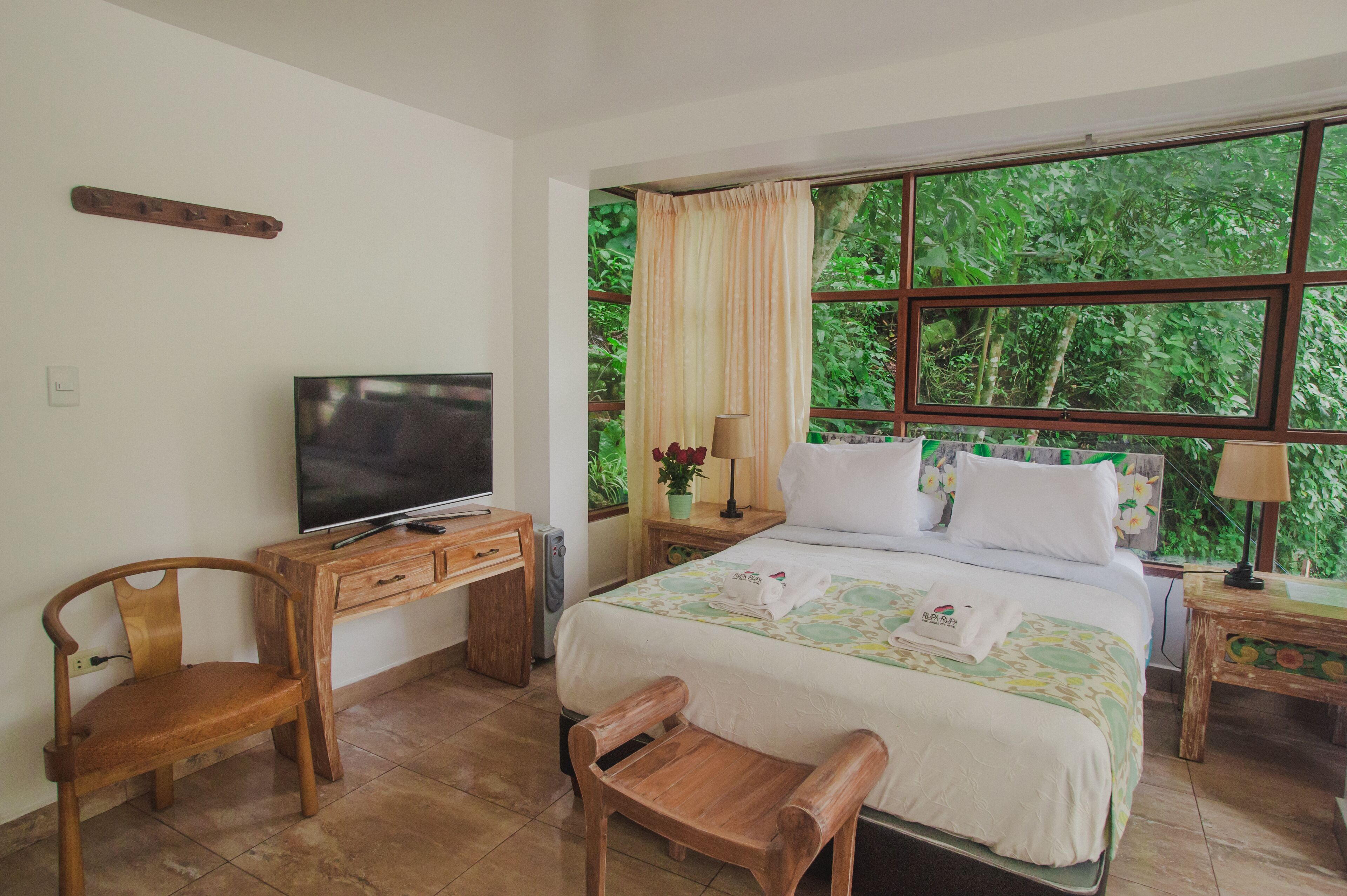 Un dormitorio con cama, televisor, muebles de madera, ventana con cortinas y vista a zonas verdes.