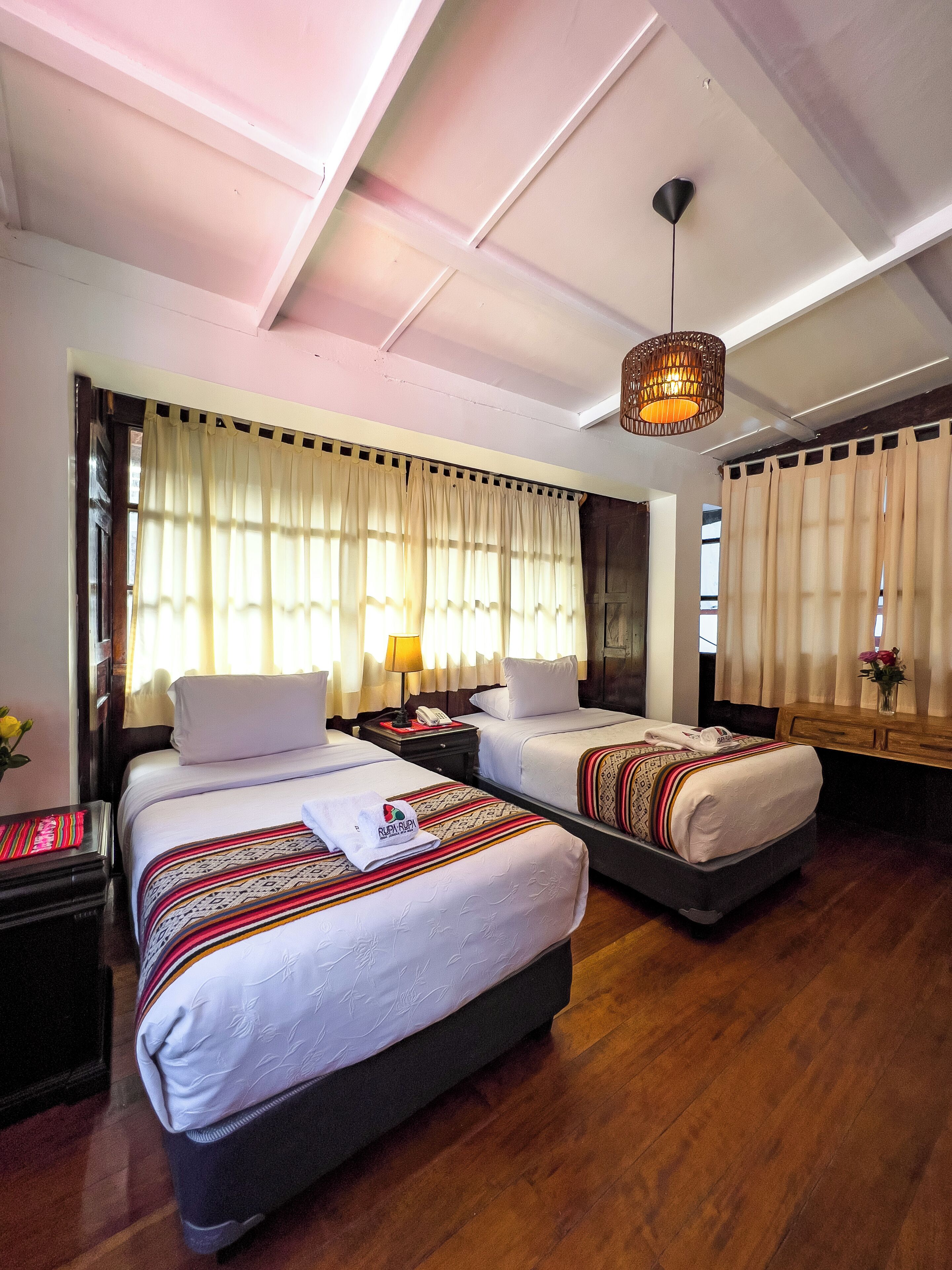 A hotel room with two beds, a wooden floor, a ceiling light fixture, and a window with curtains.