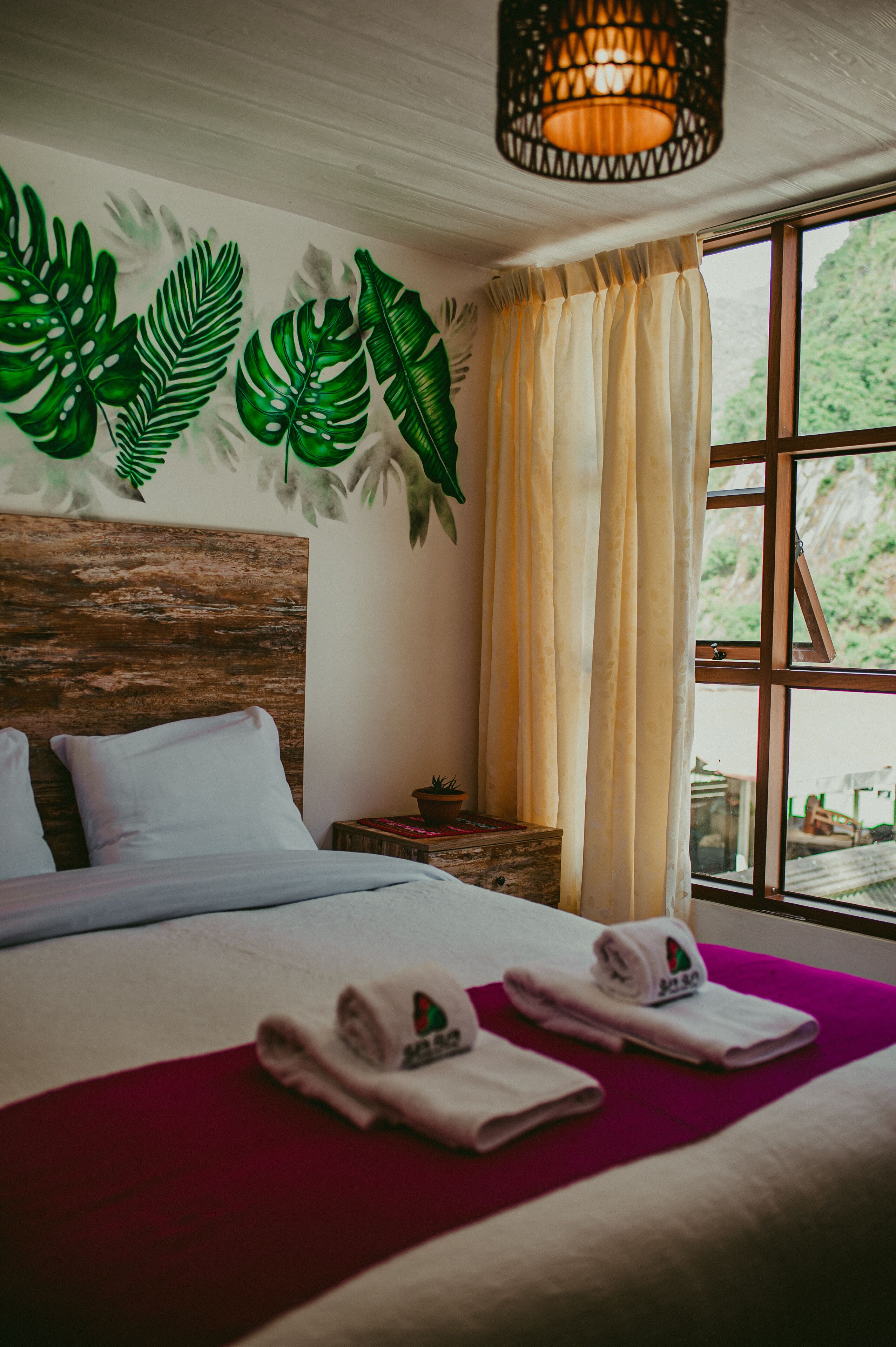 A hotel room with a wooden headboard, a bed with white linens, a red throw blanket, and a window with curtains.