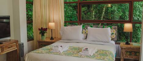 A hotel room with a bed, two bedside tables, a TV, and a window with a view of greenery.