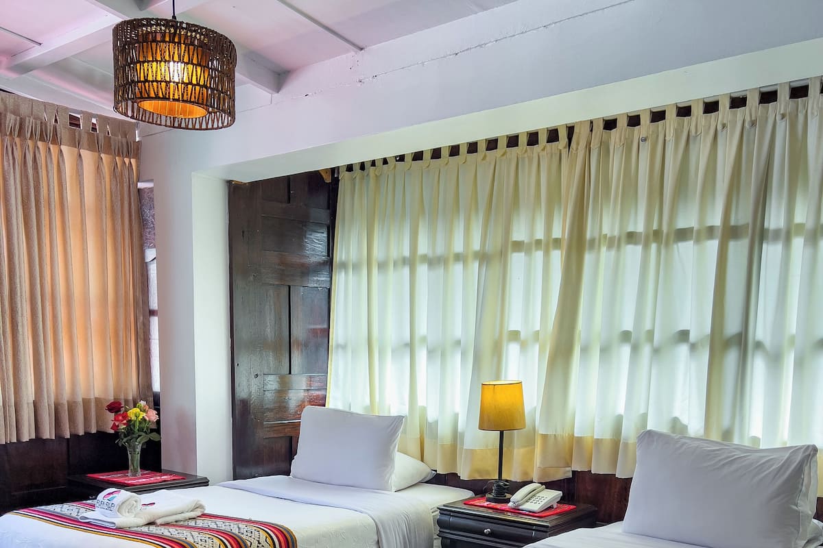 A hotel room with two beds, a wooden floor, a hanging light fixture, and a window with curtains.