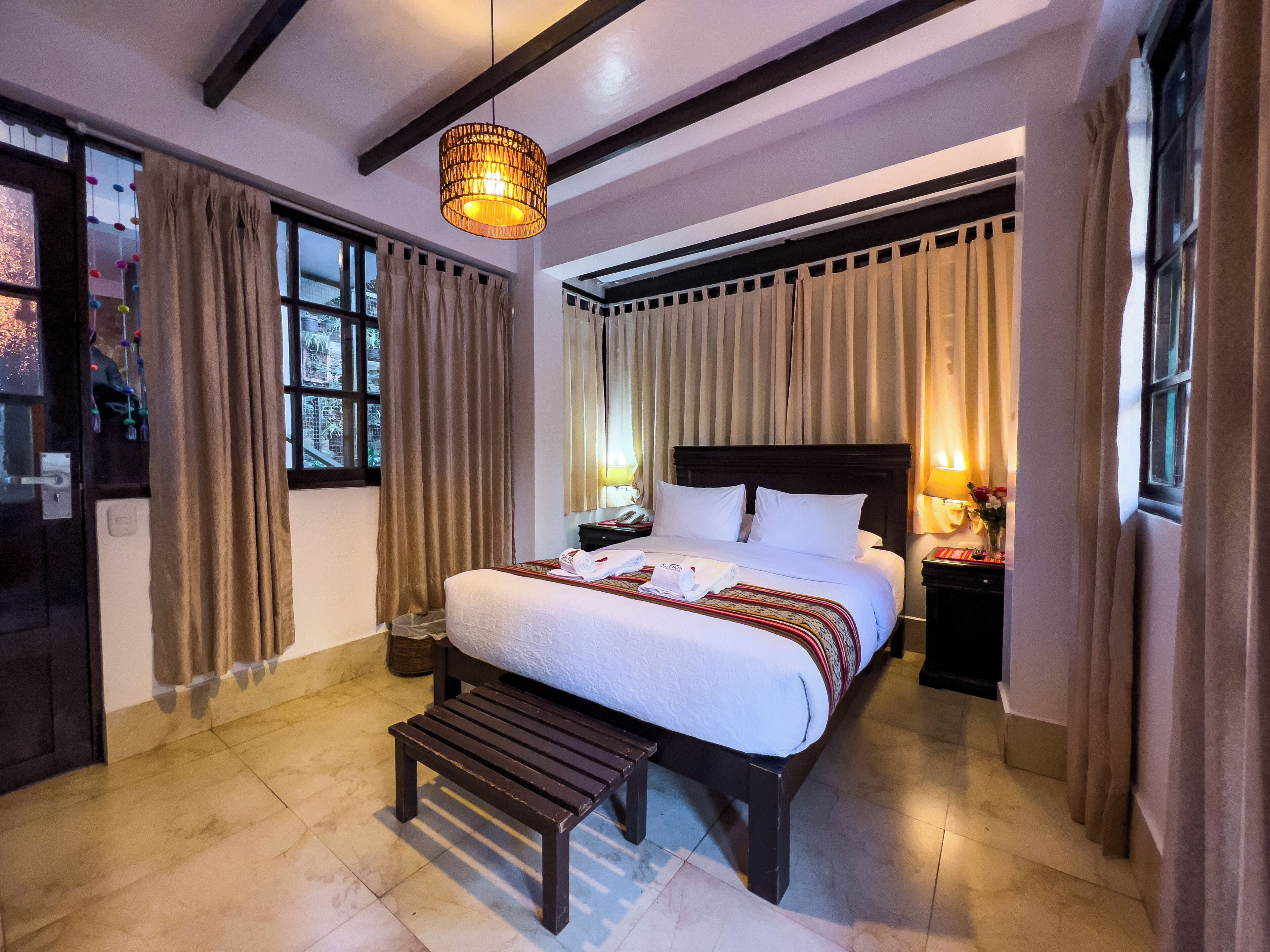 A hotel room with a large bed, a wooden bench, a hanging light fixture, and curtains.