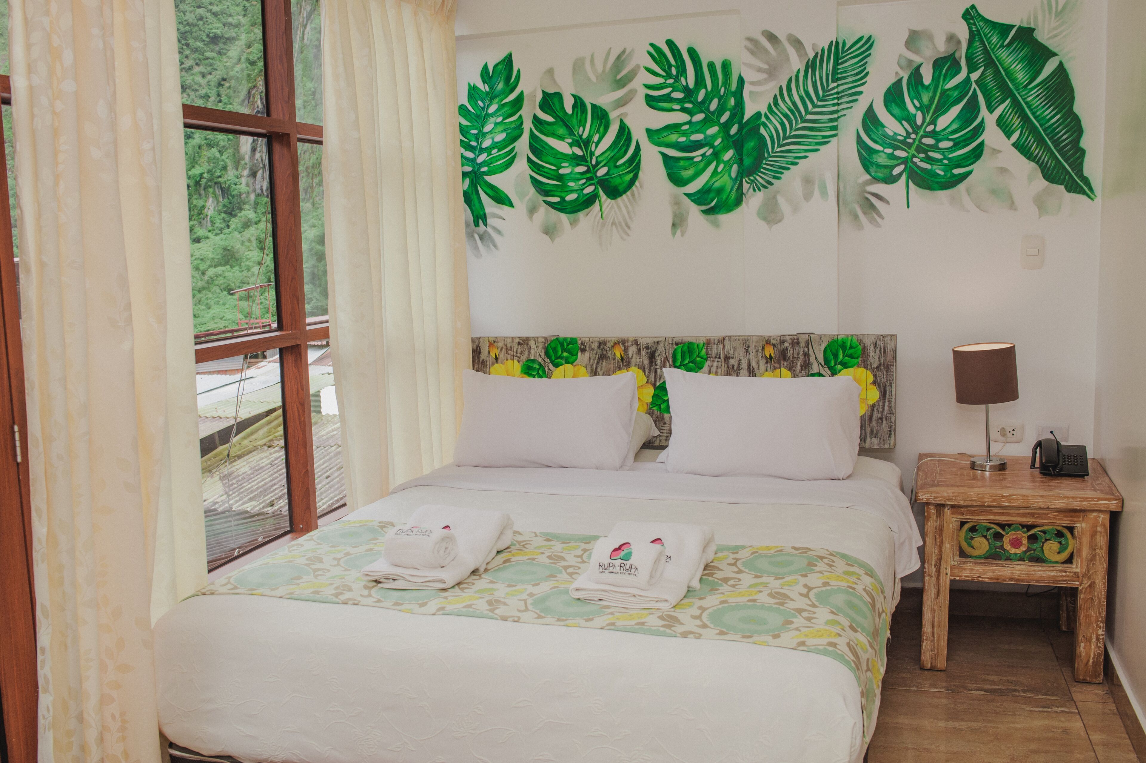 Una cama bien hecha con sábanas blancas y dos toallas, una mesita de noche con una lámpara y una pared decorada con motivos de hojas verdes.