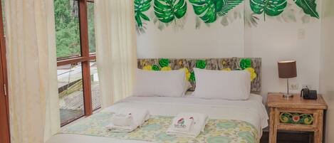 Una cama bien hecha con sábanas blancas y dos toallas, una mesita de noche con una lámpara y una pared decorada con motivos de hojas verdes.